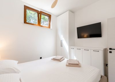 Soba bijelih zidova i bijele plahte na pruge na kojoj su četiri ručnika, bijeli ormarić iznad kojeg je plazma TV u našem šarmantnom apartmanu Lumbarda