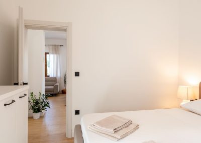 Soba s bijelim zidovima, bijelim plahtama na pruge, upaljena noćna lampa, ormarić i pogled na dnevni kroz otvorena vrata u našem šarmantnom apartmanu Lumbarda