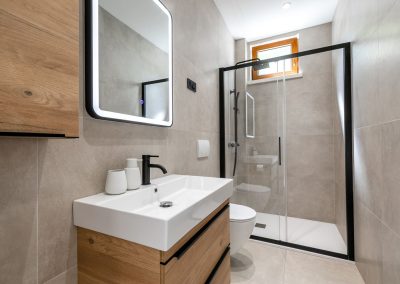 Toalet s crnim okvirom i staklom, crni tuš, bijeli umivaonik na drvenom postolju s ladicom, ogledalo s osvjetljenim okvirom u našem šarmantnom apartmanu Lumbarda