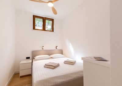Bijela soba s bijelim navlakama na pruge, noćni ormarić, prozor iznad kreveta u našem šarmantnom apartmanu Lumbarda