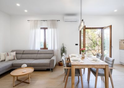 Stol za objed posložen za četvero ljudi s crvenim ružama u vazi, sivi kauč s jastucima i stolić s lampom te otvorena vrata na terasu gdje se vidi drvo i zelenilo u našem šarmantnom apartmanu lumbarda