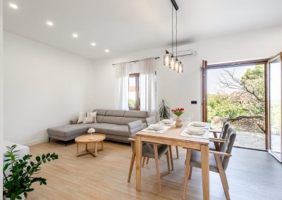 Stol za objed posložen za četvero ljudi s crvenim ružama u vazi, sivi kauč i stolić s lampom te otvorena vrata na terasu gdje se vidi drvo i zelenilo u našem šarmantnom apartmanu lumbarda